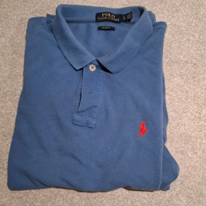 Blå pikétröja från Ralph Lauren - Säljer en klassisk blå pikétröja från Ralph Lauren med röd broderad logga på bröstet. Tröjan har korta ärmar, krage och knäppning framtill. Perfekt för en stilren och avslappnad look.