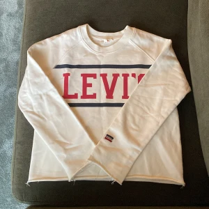 Vit långärmad tröja från Levi's - Säljer en vit långärmad tröja från Levi's med stort rött logotryck på bröstet och en liten logolapp på ärmen. Tröjan har rund halsringning och raka kanter. Perfekt för en avslappnad stil.