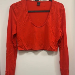 Röd croppad långärmad topp  - Säljer en röd croppad topp med lång ärm och djup rundad halsringning. Perfekt för att styla med höga jeans eller kjol. Enkel och stilren design som passar till många tillfällen.