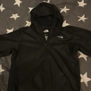 Svart vindjacka från The North Face - Säljer en svart vindjacka från The North Face med huva och dragkedja. Jackan har meshfoder på insidan och en stilren design med logga på bröstet. Perfekt för blåsiga dagar och enkel att matcha med det mesta.