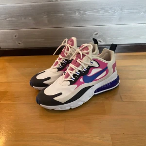 Nike Air Max 270 React sneakers i vitt och rosa - Nike Air Max 270 React sneakers med vit bas, rosa och blå detaljer samt blå sula. Skorna har snörning och en synlig Air Max-enhet i hälen för extra komfort. Perfekta för dig som gillar färgstarka och sportiga sneakers.