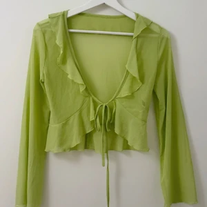 Ljusgrön volangtopp med knyt från Sahar - Trendig ljusgrön topp från Sahar med volangdetaljer och knyt framtill. Modellen är croppad med lång ärm och djup v-ringning. 