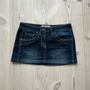 Blå lågmidjad jeanskjol  - Mörkblå lågmidjad jeanskjol med vita detaljer i storlek 34 från John Baner. Tvärs över midjan är det 38 cm och den är 29 cm lång. Använd gärna köp nu!