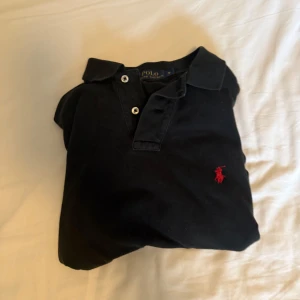 Svart pikétröja från Polo Ralph Lauren - Klassisk svart pikétröja från Polo Ralph Lauren med röd broderad logga på bröstet. Tröjan har korta ärmar, krage och knappar framtill. Perfekt för en stilren och avslappnad look. Helt ny och köptes från Johnells. Nypris 1.2 