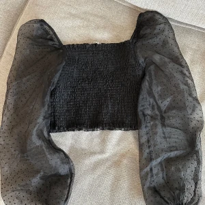 Svart croppad topp med puffärmar från NA-KD - Trendig svart  topp från NA-KD med smockad kropp och stora transparenta puffärmar i mesh med diskreta prickar. Perfekt för dig som vill sticka ut med en cool och modern look.