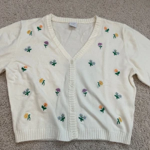 Vit kofta med broderade blommor - Söt vit kofta med broderade små blommor i olika färger. Koftan har v-ringning, knappar framtill och är croppad i modellen. Perfekt för att liva upp en enkel outfit med lite färg och mönster.