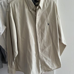 Beige skjorta från Ralph Lauren - Säljer en klassisk beige skjorta från Ralph Lauren med lång ärm och knappar framtill. Skjortan har en broderad logotyp på bröstet och är tillverkad i bomull. Perfekt för en stilren look.