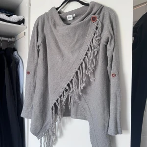 Grå stickad kofta från VILA - Säljer en grå stickad kofta från VILA i storlek XS. Koftan har asymmetrisk omlottknäppning med stora knappar, fransdetaljer längs kanten och kan bäras öppen eller stängd. Ärmarna kan vikas upp och fästas med knapp. Aldrig använd! ❤️ Önskar du veta exakta mått? Tveka inte att höra av dig! 