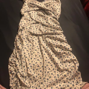 Vit blommig omlottklänning med smala axelband - Söt vit klänning med små blå blommor och smala axelband. Omlottmodell med knyt i midjan och lätt rynkade sidor. Perfekt för varma dagar och har en feminin, somrig känsla.