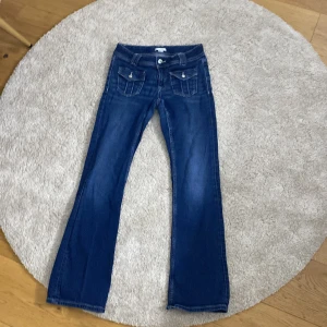 Blå bootcut jeans från Gina Tricot - Säljer ett par klassiska blå jeans från Gina Tricot i modellen bootcut. Jeansen har framfickor med lock och knappar samt markerade sömmar.
