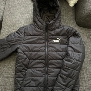 Svart pufferjacka från Puma - Säljer en svart pufferjacka från Puma med huva och dragkedja. Jackan har ett stilrent quiltat mönster och Puma-logga både fram och bak. Perfekt för kyligare dagar och enkel att matcha med det mesta. Endast testad annars är den helt ny och passar både män och kvinnor🤩.       Ordpris:629