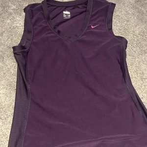 Lila ärmlös träningstopp från Nike - Säljer en lila ärmlös träningstopp från Nike med rosa logga på bröstet. Toppen har v-ringning och meshdetaljer längs sidorna för extra ventilation. Perfekt för träning och sportiga aktiviteter.