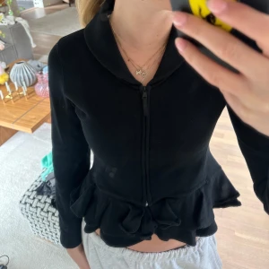 Svart volangkofta från Ralph Lauren - Säljer en svart kofta från Ralph Lauren med dragkedja framtill och volangdetalj nedtill. Koftan har lång ärm och en mjuk känsla. Snygg krage och figurnära passform som ger en feminin siluett.