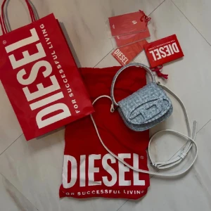 Ljusblå axelväska från Diesel - Trendig ljusblå axelväska från Diesel i jeansmaterial med vit axelrem och handtag. Väskan har ett stort D-logo framtill och stängs med lock. Perfekt för att bära det viktigaste och ge din outfit en cool touch.