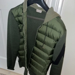 Grön Jacka med huva från Moncler - Snygg grön jacka från Moncler med stickade ärmar och rygg samt quiltad framsida. Jackan har huva, dragkedja och en stor svart detalj på ärmen. Perfekt för dig som vill ha något unikt och stilrent. Moncler. Storlek M. Använd 2 gånger.
