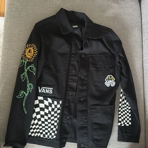 Svart overshirt från Vans med tryck - Säljer en svart overshirt från Vans i storlek XS. Jackan har coola tryck med solros, svamp och schackrutor samt texten 'Off The Wall' på ryggen. Flera fickor framtill och knäppning med knappar. Perfekt för dig som gillar streetwear.
