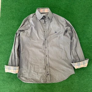 Grå skjorta från Burberry med rutiga detaljer - Stilren grå skjorta från Burberry i bomull, med klassiskt rutigt mönster på insidan av krage och ärmslut. Skjortan har knappar framtill och lång ärm, perfekt för en dressad look.