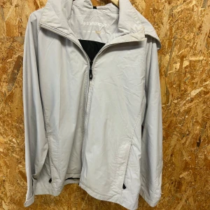 Ljusgrå vindjacka från Everest - Snygg ljusgrå vindjacka från Everest med huva och dragkedja framtill. Jackan har två fickor med dragkedja och en enkel, stilren design. Perfekt för blåsiga dagar och enkel att matcha med olika outfits.