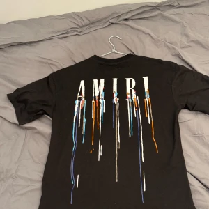 Svart t-shirt från Amiri med färgstänk - Säljer en svart t-shirt från Amiri med tryckt logga och färgstänk i blått, vitt och orange på både bröstet och ryggen. T-shirten har rund hals och normal passform. Perfekt för dig som gillar streetwear och unika detaljer. Den är använd fåtal gånger och är i jätte bra skick tag fås med, fraktas direkt vid köp 
