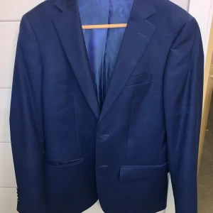 Mörkblå kostym med kavaj och byxor - Säljer en stilren mörkblå kostym med matchande kavaj och byxor. Kavajen har klassisk passform, två knappar framtill och fickor med lock. Perfekt för formella tillfällen som bal eller student.