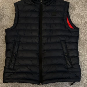 HUGO BOSS Väst  - Säljer en mörkblå dunväst från BOSS som är i storleken M/S. Västen har två fickor med knappar och en snygg röd insida som ger en cool kontrast. Perfekt för lager-på-lager och stilren look.