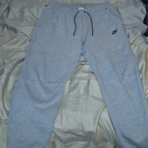 Grå mjukisbyxor från Nike tech - Säljer ett par grå mjukisbyxor från Nike med svart logga på vänster ben. Byxorna har dragsko i midjan och två sidofickor. Perfekta för chill eller träning, klassisk look med bekväm passform. STORLEK. L