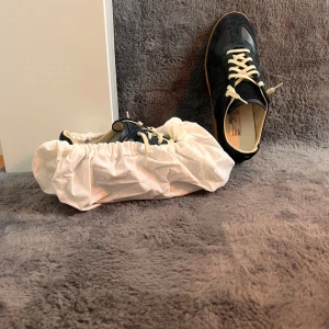 Svarta sneakers från Maison Margiela - Säljer ett par svarta sneakers från Maison Margiela med beige snörning och gummisula. Skorna har en klassisk design med rund tå och detaljer i mocka och skinn. Perfekta för dig som gillar stilrena och exklusiva sneakers.