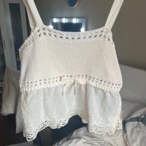 Vit stickad och spetsprydd linnetopp - Söt vit linnetopp med stickad överdel och breda axelband. Nedre delen är i tunt tyg med broderade spetsdetaljer och liten rosett framtill. Perfekt för varma dagar och ger en romantisk känsla.