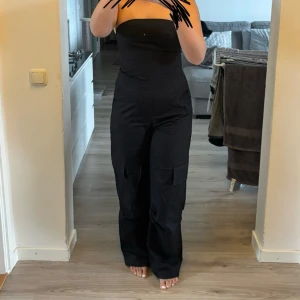 Svart byxdress med cargofickor - Säljer en svart byxdress från Zara med raka ben och cargofickor på sidorna. Byxdressen är axelbandslös och har en figurnära passform upptill. Perfekt för dig som vill ha en stilren och modern look.