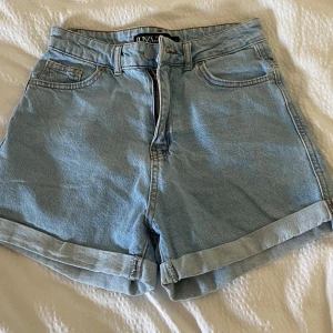 Ljusblå jeansshorts med hög midja - Säljer ett par ljusblå jeansshorts från Monki med hög midja och klassisk femficksdesign. Shortsen har uppvikta benslut och stängs med dragkedja och knapp. Perfekta för varma dagar!