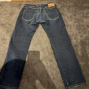 Mörkblå Replay Anbass jeans - Säljer ett par mörkblå Replay Anbass jeans med klassisk femficksdesign och raka ben. Jeansen har snygga kontrastsömmar och läderpatch bak i midjan. Perfekta för dig som gillar stilrena och tidlösa jeans.