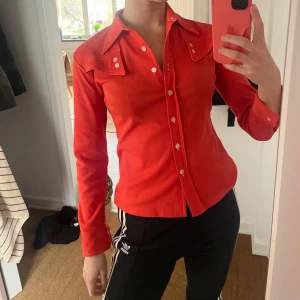 Röd retro skjorta med vita knappar - Snygg röd/orange skjorta i retrostil med långa ärmar och markerad krage. Skjortan har vita knappar framtill och på bröstfickorna samt dekorativa sömmar. Tillverkad i 100% polyester, perfekt för dig som gillar färg och vintage vibe. Köpt här på plick och bara använd någon gång! Lite cowboy vibe 🐄🤠