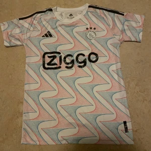 Ajax fotbollströja  - Säljer en Ajax fotbollströja från Adidas med korta ärmar. Tröjan har ett unikt mönster i vitt, blått och rött med svarta detaljer och Ziggo-logga på bröstet. Materialet är lätt och andas, perfekt för träning eller match.