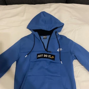 Blå hoodie från Nike - Säljer en blå hoodie från Nike med 'Just Do It' tryck på framsidan. Tröjan har en dragkedja vid halsen och en stor ficka framtill. Perfekt för en sportig look!