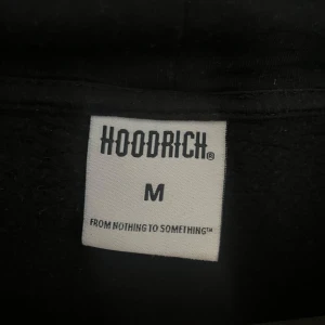Svart hoodie från Hoodrich - Säljer en svart hoodie från Hoodrich med stor logga på både fram- och baksidan. Hoodien har en klassisk känguruficka och justerbar huva med dragsko. Perfekt för en avslappnad stil. Den har en liten ficka på undersidan av armen.