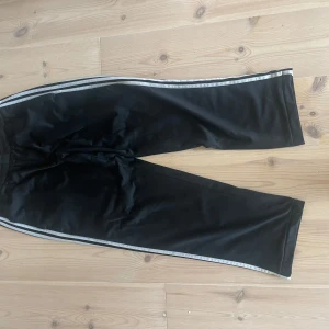 Svarta träningsbyxor från Adidas - Säljer ett par svarta träningsbyxor från Adidas med vita ränder längs sidorna. Byxorna har en elastisk midja och är perfekta för träning. De är tillverkade i ett syntetmaterial som ger bra rörlighet.