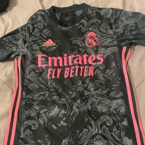 Svart och grå Real Madrid fotbollströja - Snygg svart och grå fotbollströja från Adidas med Real Madrid-logga och rosa detaljer. Tröjan har ett unikt mönster och korta ärmar. Perfekt för alla fotbollsfans som vill sticka ut på planen.