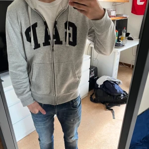 Grå hoodie från GAP - Säljer en  grå hoodie från GAP med dragkedja och stor logga på bröstet. Tröjan har en bekväm passform i storlek M. Hör av dig vid fler frågor🙏🏼