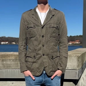 Olivgrön fieldjacket - Snygg olivgrön tunn fieldjacket med knappar och flera fickor. Perfekt för en varmare vårdag!