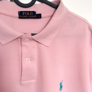 Rosa pikétröja från Ralph Lauren - Säljer en stilren rosa pikétröja från Ralph Lauren med klassisk krage och knappar framtill. Tröjan har en broderad blå logotyp på bröstet. Perfekt för en avslappnad och snygg look. Storlek L, och endast använd ett fåtal gånger i sommar.