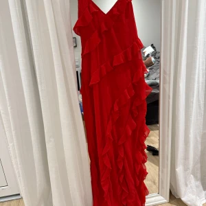 Röd långklänning med volanger - Säljer en fantastisk röd långklänning med volanger. Klänningen har en v-ringad design och är perfekt för festliga tillfällen. Den är ärmlös och har en elegant och flytande passform.