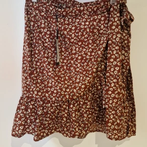 Blommig omlottkjol från Only - Snygg blommig kjol från Only i en härlig brun färg med vita blommor. Kjolen har en volangdetalj längst ner och är tillverkad i 100% polyester. Perfekt för en somrig look!