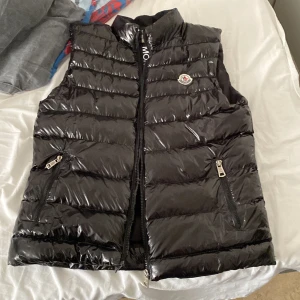 Moncler väst  - Snygg svart Moncler väst med glansig finish. Västen har dragkedja framtill och två praktiska fickor inklusivt i insidan med dragkedjor. Utttråkad av den nu har haft den i 6 månader ungefär. Storlek S men kan passa M.