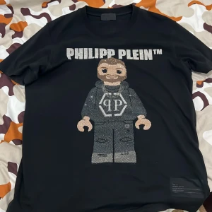 Svart t-shirt från Philipp Plein med glittrig design - Säljer en svart t-shirt från Philipp Plein med en cool glittrig figur på framsidan. T-shirten har korta ärmar och är gjord i bomull. Perfekt för dig som vill sticka ut med stil!