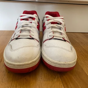 New Balance 550 sneakers i vitt och rött - Snygga New Balance 550 sneakers i vitt med röda och svarta detaljer. Skorna har snörning och en rund tå, vilket ger en klassisk look. Perfekta för en sportig stil.