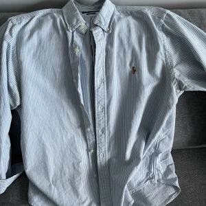 Randig skjorta från Polo Ralph Lauren - Säljer en randig skjorta från Polo Ralph Lauren i blått och vitt. Storlek L. Skjortan är använd 3-4 gånger men har hängt i garderoben ett tag eftersom den är för stor för mig. Priset är väldigt lågt då jag bara vill bli av med den.