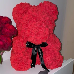 Röd nallebjörn av rosor - Charmig nallebjörn täckt av röda rosor, perfekt som en romantisk present eller dekoration. Den har ett svart band knutet runt halsen med text. En unik och söt detalj för att uttrycka kärlek eller uppskattning.