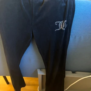Svarta mjukisbyxor från Juicy Couture - Säljer ett par svarta mjukisbyxor från Juicy Couture med en glittrig logga på ena benet. Byxorna har en bekväm passform och är perfekta för en avslappnad stil.