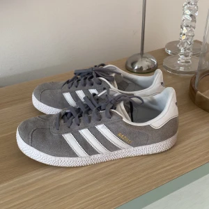 Grå Adidas Gazelle sneakers - Snygga grå Adidas Gazelle sneakers i mocka med de klassiska vita ränderna på sidan. Skorna har snörning och en vit sula som ger en stilren look. Perfekta för en avslappnad stil. nypris 1200. 