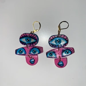Färgglada örhängen med ögonmotiv - Unika och färgglada örhängen med ögonmotiv i blått och rosa. De har en lekfull design med flera ögon och läppar, och är fästa med guldfärgade krokar. Perfekta för att ge en konstnärlig touch till din stil.
